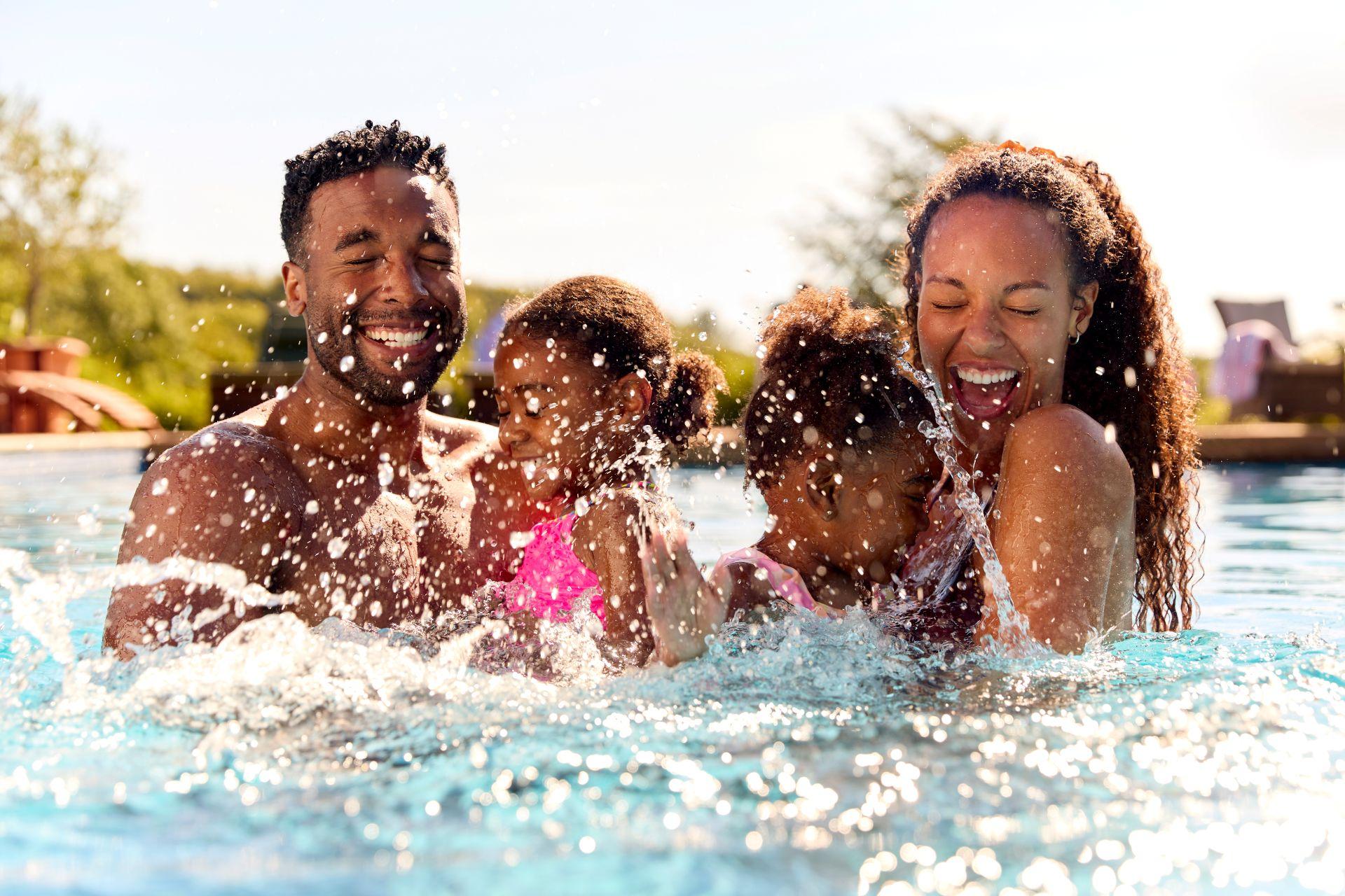 Familie mit zwei Kindern spielt lachend im Pool, Wasserspritzer in der Sonne